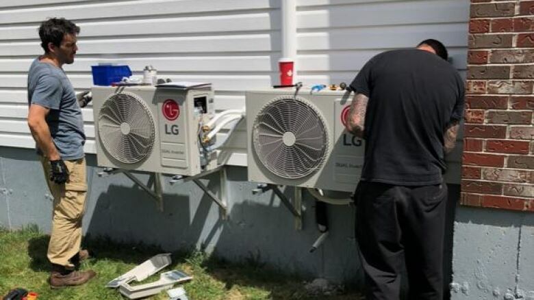 Two men at work installing two exterior heat pump units on the side of a house with some wire and tools on the lawn in the foreground