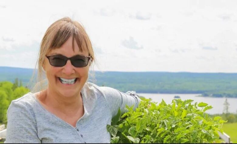 A woman with light brown hair, wearing sunglasses, smiling broadly and holding a basket of greens, with a view of a river valley in the background