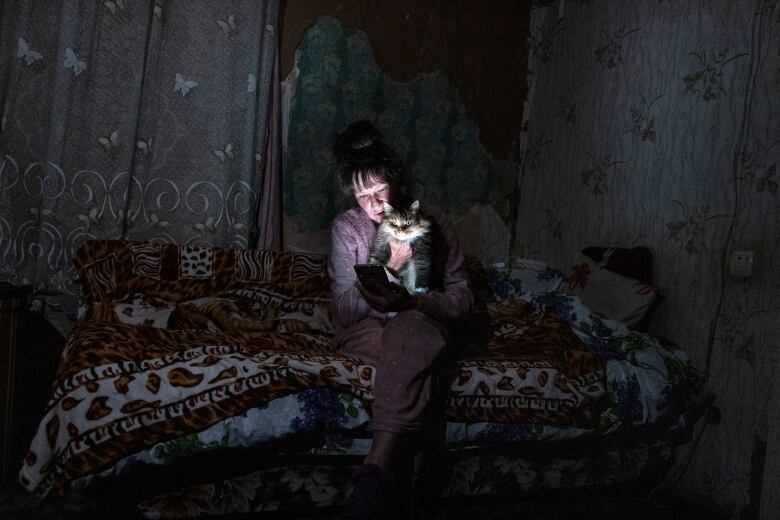 A woman sits on a bed in a dark room in her pajamas snuggling a cat, lit only by the light of her phone. 