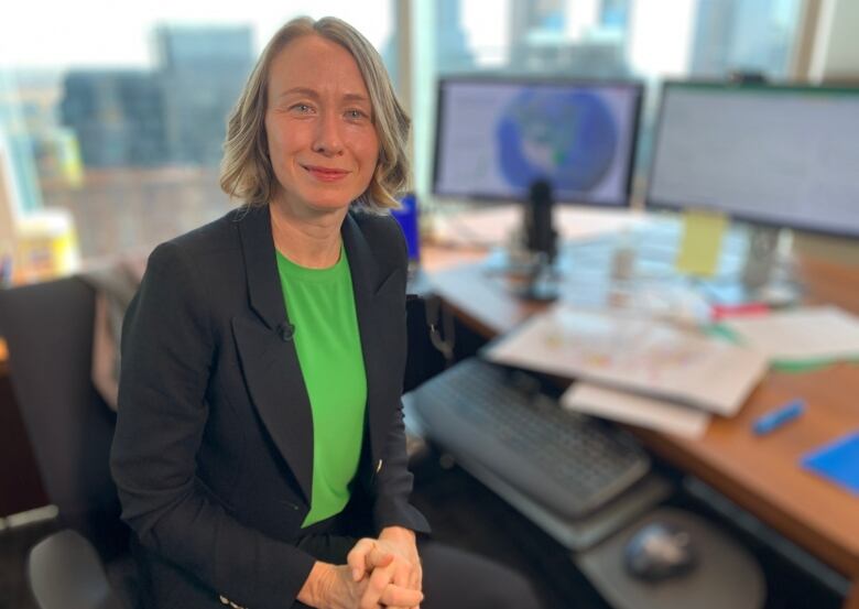 A woman with short blond hair and wearing a business suit sits at a desk with computer monitors.