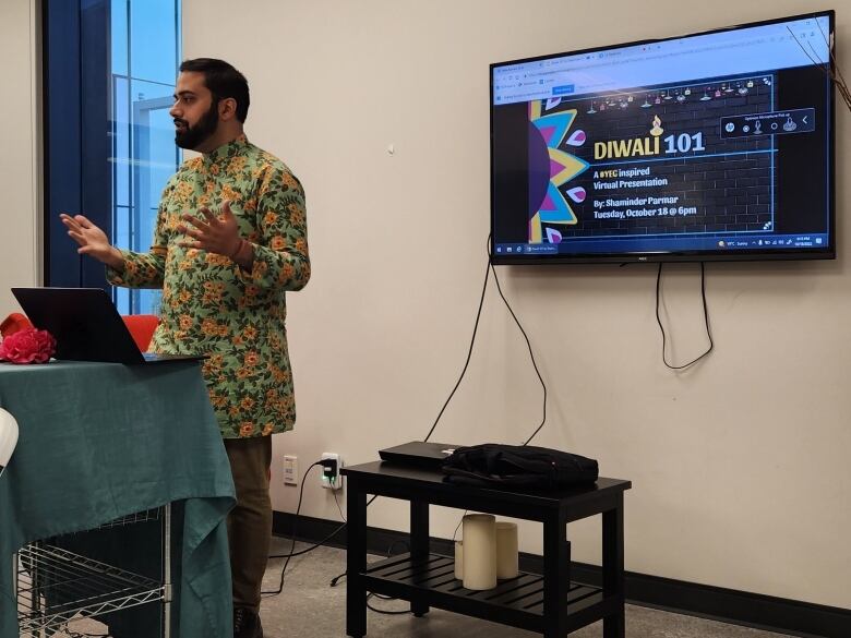 A man stands wearing a long sleeved printed floral tunic. He's on a laptop presenting out to an audience that's behind the camera. Behind him a TV screen is playing a powerpoint presentation, with a slide that says Diwali 101.