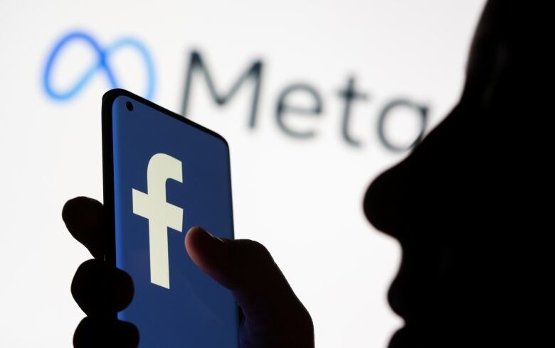 A silhouetted person holds a phone with the Facebook logo on it.