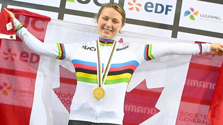 A woman with blond hair wearing a gold medal around her neck holds a Canadian flag behind her with arms spread wide.