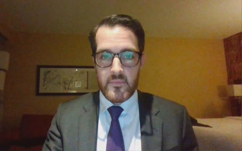 A chest-up shot of a person in a suit and wearing glasses sitting in a room with low lighting.