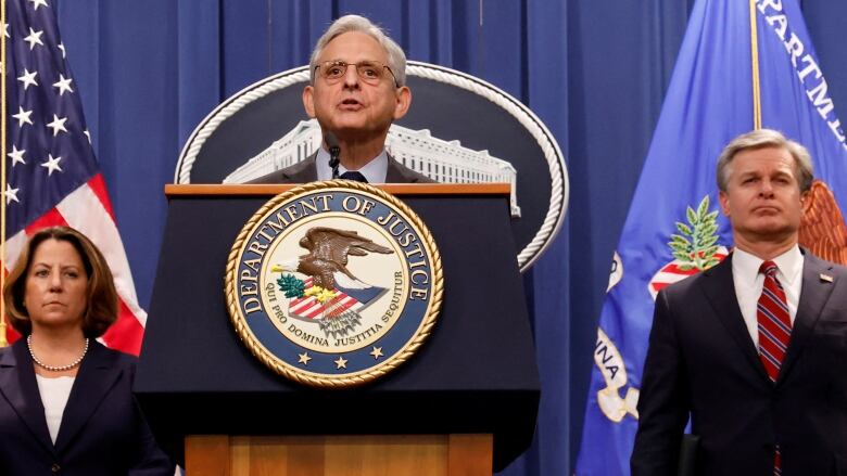 A grey haired individual wearing a blue suit stands at a podium with a Department of Justice flag on the front.