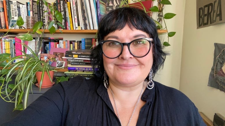 A selfie photo of a person wearing a black dress shirt, black-rimmed glasses and silver jewellery, in front of a wall with books and plants on shelves.