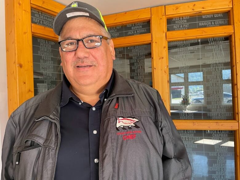 A First Nations chief in glasses and a baseball hat wears a black jacket with his band's name and the word chief embroidered on it. 