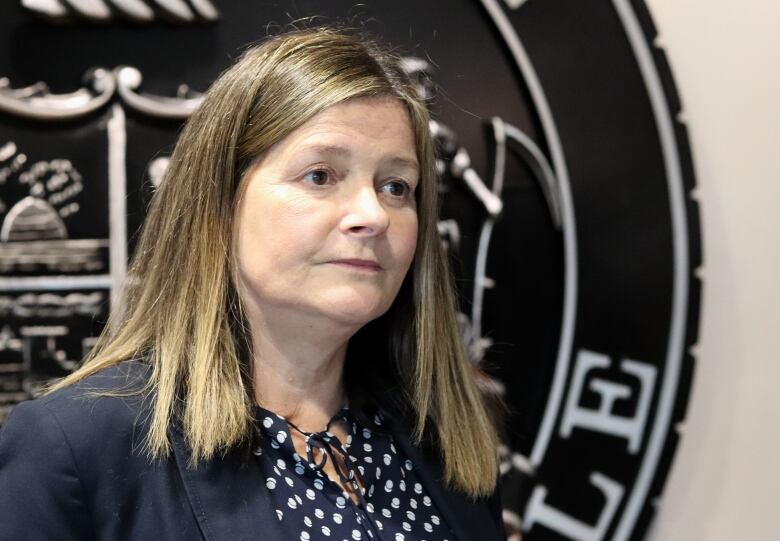 A woman with shoulder-length brown hair wearing a black jacket over a polka-dot shirt in front of a black metallic City of Moncton logo.