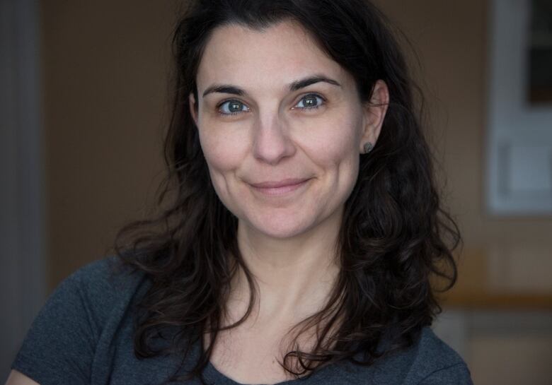 A woman with below shoulder-length brown hair sits centre in front of a dark background. She wears a dark blue t-shirt.