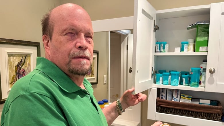 A man, standing in a bathroom, holds open the door to a medicine cabinet, revealing about a dozen bottles and packages of prescription drugs.