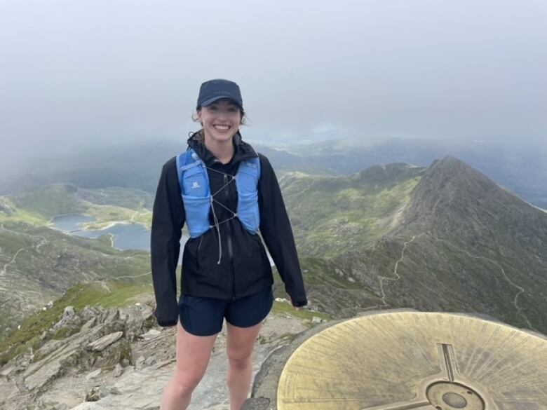 A woman stands on top of a mountain smiling at the camera.