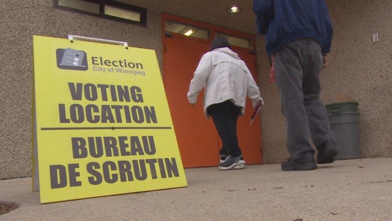 People wearing light jackets walk toward a pair of orange doors. A sign in the foreground says 