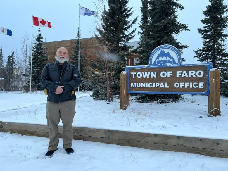 A man with a white beard stands outside next to a sign for the Town of Faro Municipal Office.