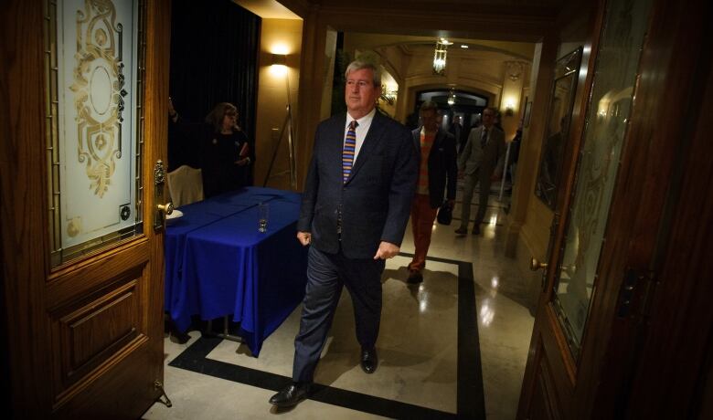 A man in a suit looks sad as he walks down a hallway. Several people trail behind him.