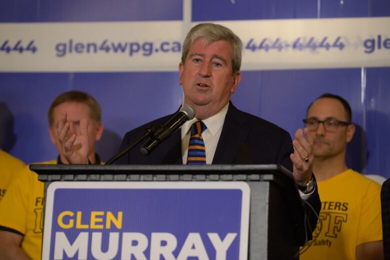 A man is standing at a podium speaking into a microphone, with a sign that says 