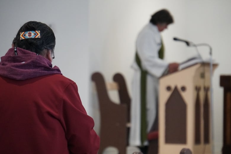 A person sits with their back to the camera facing a minister who is at a podium speaking.