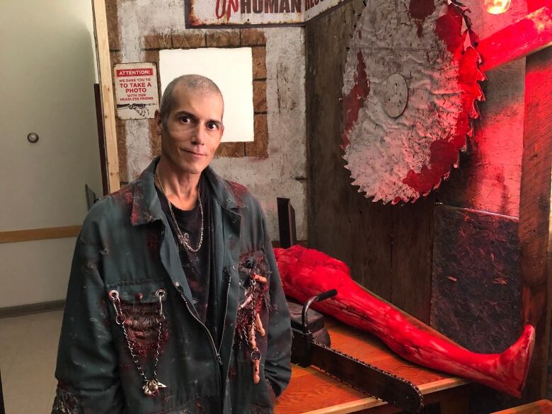 A bald man in costume with a bloody jacket and props of a buzz saw and fake body in the background.