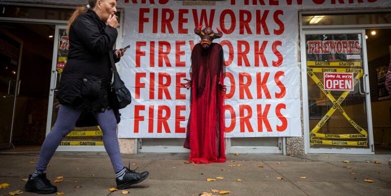 A woman walks past a store with numerous signs advertising fireworks, plus a guy in a devil costume