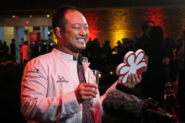 A smiling man in a white jacket holds a glass of champagne and a white star symbolizing his restaurant's entry to the Michelin Guide.