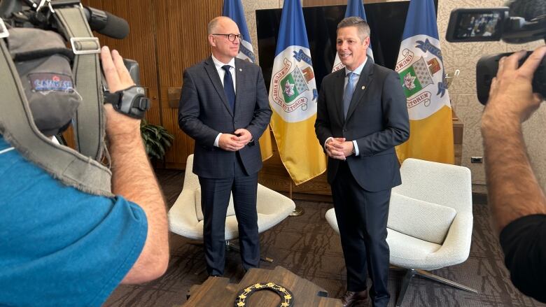 Two men wearing suits are standing next to each other in an office with television cameras pointed at them.