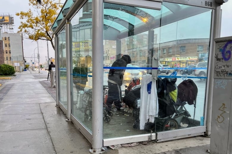 Several people gather in a bus shelter with glass walls on a busy city street.