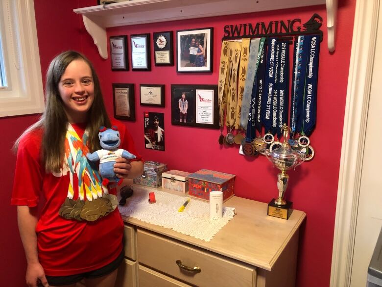 A woman wearing swimming medals holding a teddy bear standing beside a wall featuring some of her swimming accomplishments