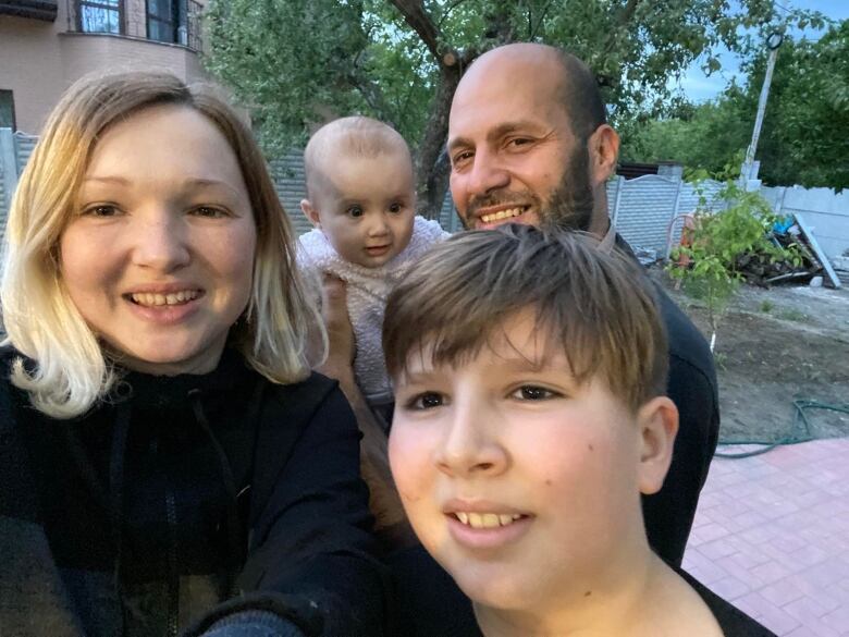 A woman stands beside a teenage boy and a man holds a baby in the background. They are all smiling. 