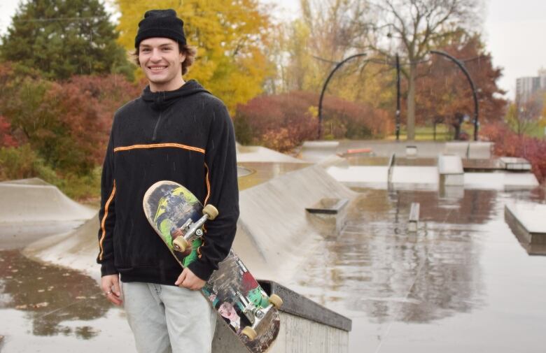 Young man wearing a tuque (or beanie) holding a skateboard. The skatepark has concrete ramps. Trees with leaves that have changed colours are seen in the background and the concrete is wet because it was raining.