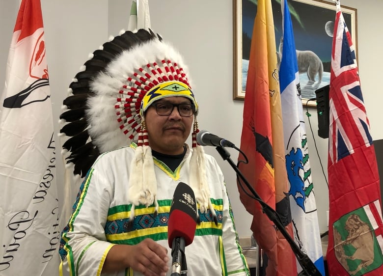 A man wearing an Indigenous war bonnet speaks at a podium.