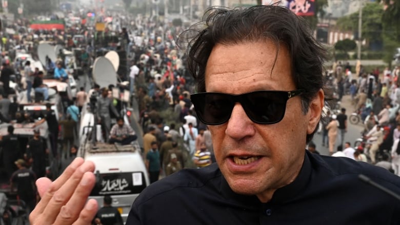 Imran Khan wears dark glasses and a dark jacket. He gestures in front of a busy scene.