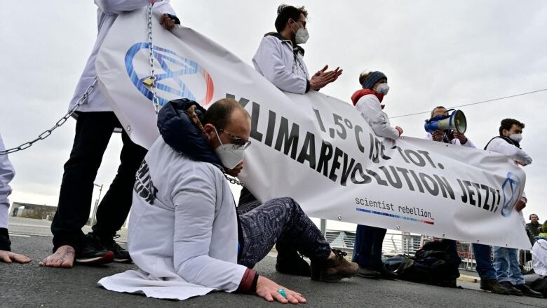 A group of people wearing white jackets and masks hold up a massive banner. One man in the front is on the ground with his palm flat on the ground alongside him. 