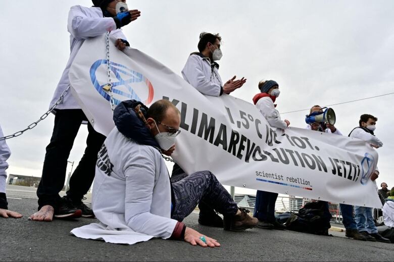 A group of people wearing white jackets and masks hold up a massive banner. One man in the front is on the ground with his palm flat on the ground alongside him. 