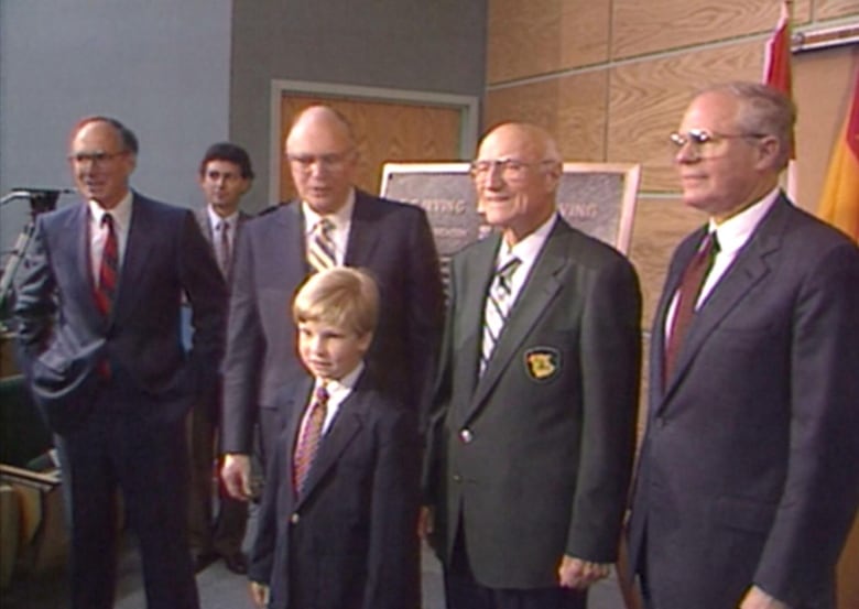 Four men in business suits stand together in a row. In front of them is a boy, also dressed in a suit. 