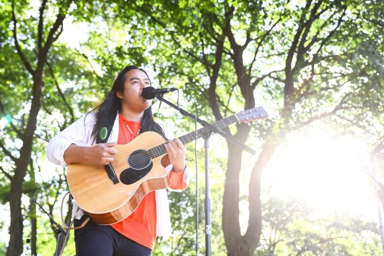 A man plays a guitar and sings in front of a microphone outside in a park.