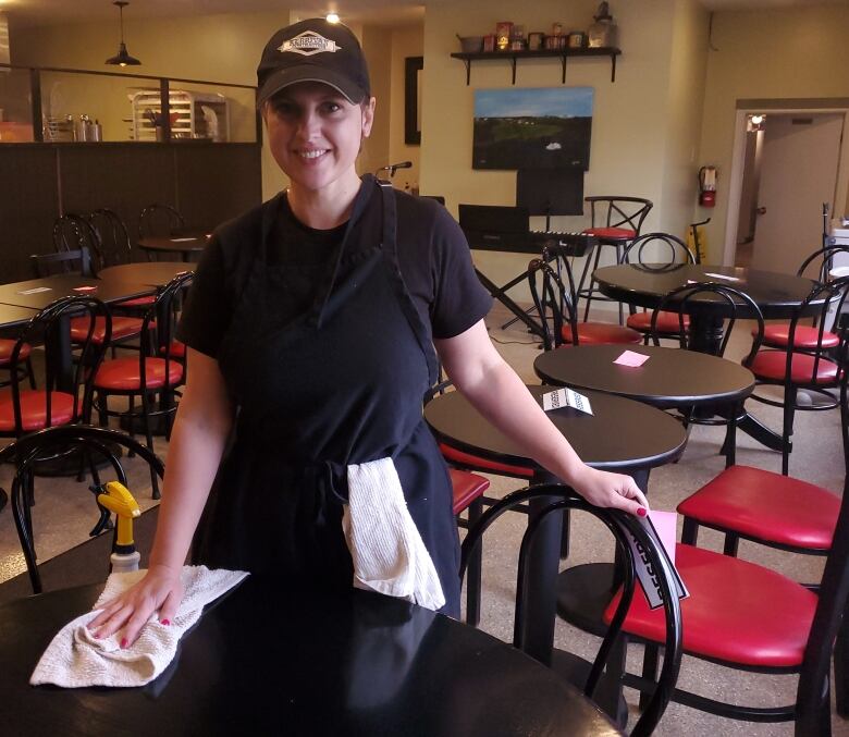 A young woman wearing a black baseball hat and black uniform cleans a table while working at a cafe.