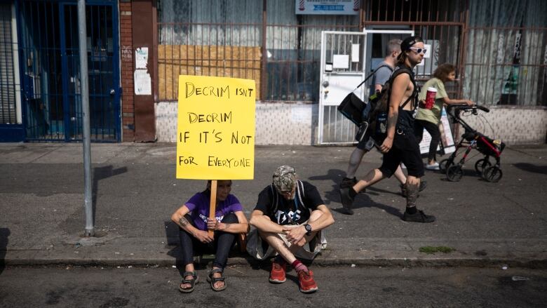 Two people sitting on the sidewalk hold up a sign that reads 'Decrim isn't decrim if it's not for everyone'.