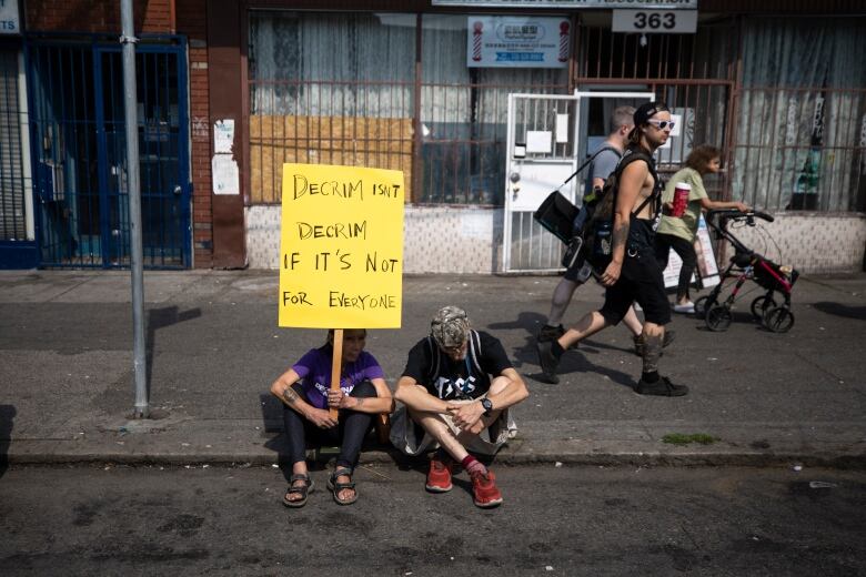 Two people sitting on the sidewalk hold up a sign that reads 'Decrim isn't decrim if it's not for everyone'.