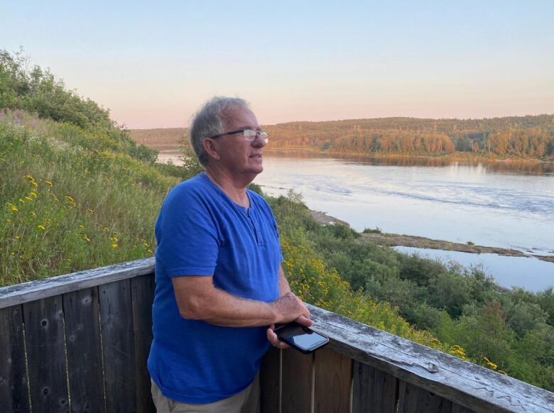 An older man in glasses and a blue T-shirt leans against a deck railing, looking out at a river.