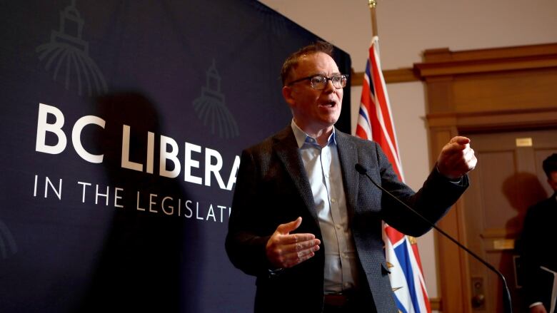 Kevin Falcon gestures at a podium. He is a white man wearing a suit and glasses.