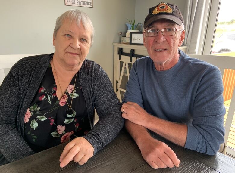 Two people sit at a table. The person on the right is wearing a shirt with flowers on it and a necklace. The person on the right wears a baseball hat. Both have grey hair.