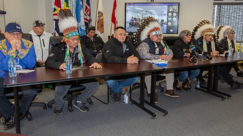 Seven people are seated at a table, including four men in headdresses, with others seated behind them.