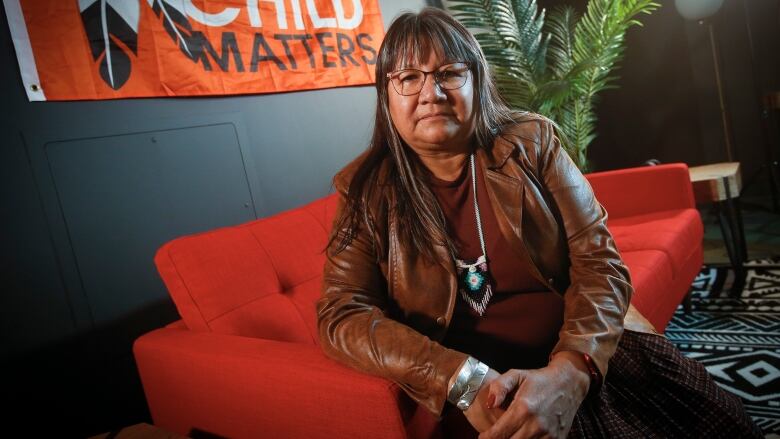 A woman with long dark hair and glasses sits on a red couch, looking at the camera, with a flag reading 