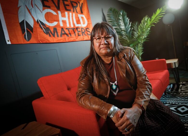 A woman with long dark hair and glasses sits on a red couch, looking at the camera, with a flag reading 