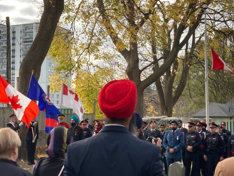 A man wearing a red turban stands outside facing away from camera. Other people in uniforms can be seen in the background during a remembrance ceremony.