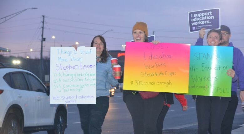 Striking education workers carry colourful signs, including one listing things the woman trusts more than Stephen Lecce, while walking in the early morning light along Barrydowne Road  