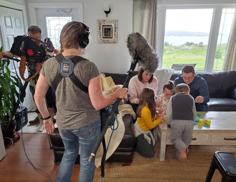 A television crew of a videographer and an audio technician record the White family. A man and woman sit on a couch as three small children play along a coffee table. 