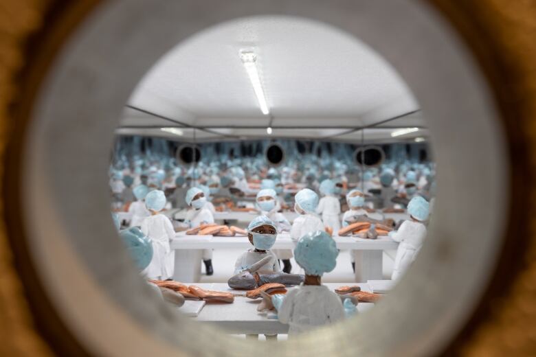 Photo of a box diorama of miniatures. The figures are factory workers in a fish processing facility. They wear smocks and blue face masks and hair nets and appear in infinite rows, cutting pink slabs of fish at white tables.