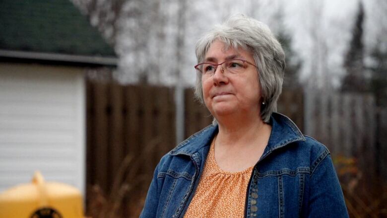 A grey-haired woman in glasses and a jean jacket stands in her backyard. 