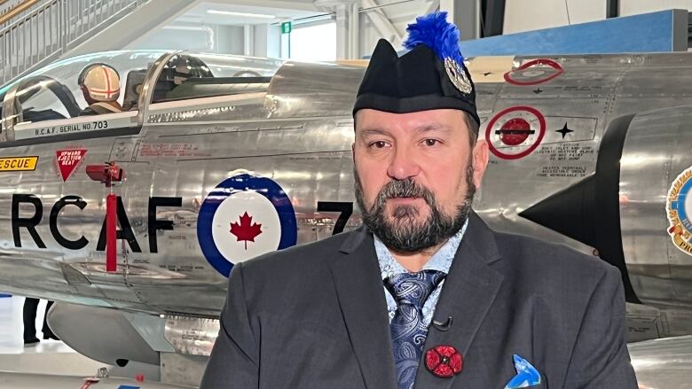 A man wearing a military cap and a blue jacket with a poppy pinned to the lapel stands in front of an airplane in a hangar.