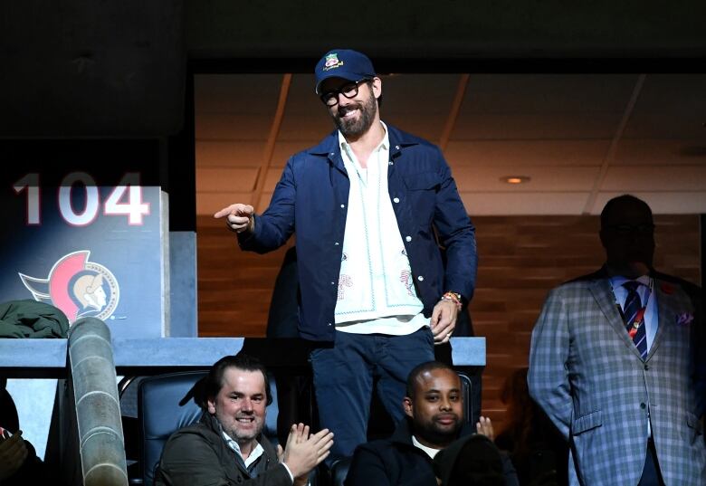 A man under a spotlight in an arena suite points at someone off-camera.